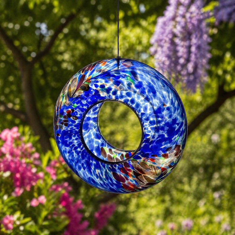 May include: A circular glass bird feeder featuring a vibrant blue speckled pattern with accents of red, green, and white. The hollow center provides an open space, and the piece is suspended by a thin black cord for hanging in an outdoor setting.