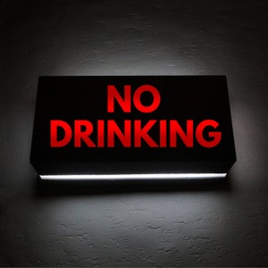 Könnte beinhalten: Ein rechteckiges schwarzes Schild mit den Worten "NO DRINKING" in leuchtend roten Buchstaben. Ein weißes Licht beleuchtet die Unterkante des Schildes und erzeugt einen Kontrast zum dunklen Hintergrund.