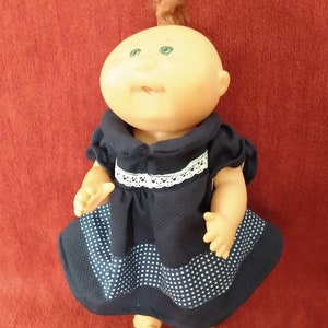 May include: A vintage doll with a bald head, green eyes, and a small tuft of brown hair. The doll is wearing a navy blue dress with white polka dots and a white lace trim.