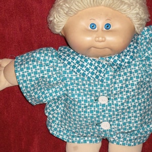 May include: A Cabbage Patch Kid doll wearing a blue and white checkered shirt with a white collar. The doll has blonde hair and blue eyes.