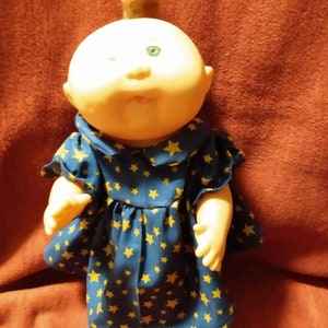 Pode incluir: Uma boneca Cabbage Patch Kid vestindo um vestido azul com estrelas amarelas. A boneca tem um rabo de cavalo loiro e olhos verdes.