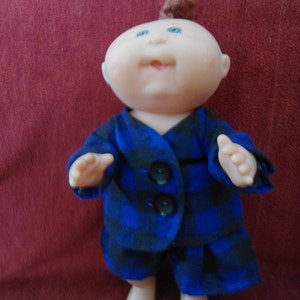Peut inclure: Une poupée Cabbage Patch Kid portant une tenue en tissu à carreaux bleus et noirs avec trois boutons noirs. La poupée a le teint rosé, les yeux bleus et une crête brune.