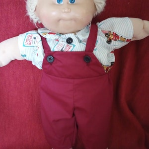 Könnte beinhalten: Eine Cabbage Patch Kid Puppe mit blonden Haaren, blauen Augen und einem rot-weißen Outfit. Die Puppe trägt ein weißes Hemd mit einem blau-roten Muster und eine rote Latzhose mit schwarzen Knöpfen. Die Puppe sitzt auf einer roten Oberfläche.