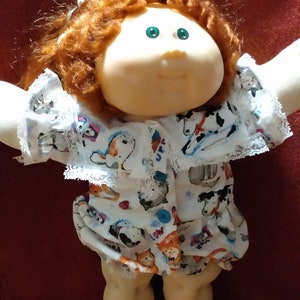 May include: A Cabbage Patch Kid doll with red curly hair wearing a white and blue patterned dress with a lace trim. The dress has a variety of animal illustrations, including cats, dogs, and horses.
