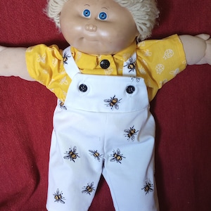 May include: A Cabbage Patch Kid doll wearing a yellow short-sleeved shirt with a bee pattern and white overalls with a bee pattern.