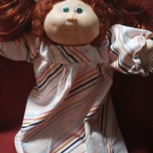 May include: A red-haired Cabbage Patch Kid doll wearing a white and pink striped dress with blue, brown, and orange stripes.