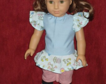 Puede incluir: Una muñeca con un vestido azul claro con una falda de estampado floral y pantalones cortos rosas. La muñeca tiene el pelo castaño y los ojos azules.