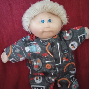 Pode incluir: Uma boneca Cabbage Patch Kid com cabelo loiro e olhos azuis vestindo um macacão cinza e laranja com tema de videogame. O macacão tem uma variedade de gráficos, incluindo controladores de jogos, fones de ouvido e as palavras "Battle Royale".