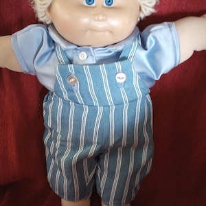 Könnte beinhalten: Eine Cabbage Patch Kid Puppe mit blonden Haaren, blauen Augen und einem blau-weiß gestreiften Outfit. Die Puppe sitzt auf einer roten Oberfläche.