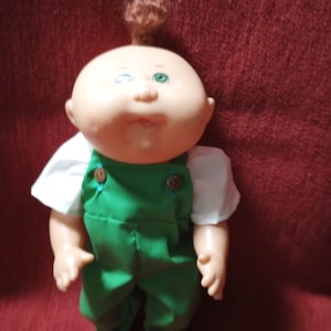 May include: A Cabbage Patch Kid doll with red hair, wearing a white shirt and green overalls. The doll has green eyes and a rosy complexion.