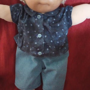 Könnte beinhalten: Eine Cabbage Patch Kid Puppe mit blonden Haaren, blauen Augen und einem blau-weiß gestreiften Hemd. Die Puppe trägt eine blaue Jeanshose und sitzt auf einer roten Oberfläche.
