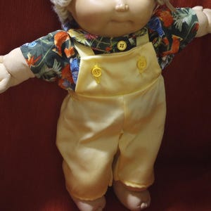 May include: A doll with light skin, blue eyes, and blonde yarn hair. The doll wears a dark green shirt featuring a colourful floral print with orange and blue flowers, paired with bright yellow overalls secured by two yellow buttons on the front.