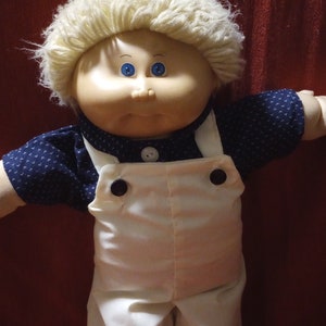 May include: A Cabbage Patch Kid doll with blonde hair, blue eyes, and a white outfit with blue accents. The doll is wearing overalls and a blue shirt with white polka dots.