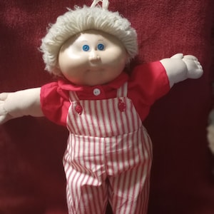 Może przedstawiać: Lalka Cabbage Patch Kid z blond włosami, niebieskimi oczami i czerwono-białą paskowaną kombinezonem.