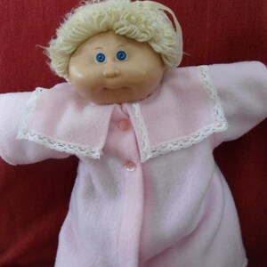 Könnte beinhalten: Eine Cabbage Patch Kid Puppe, die einen rosa Fleece-Schlafanzug mit weißer Spitzenborte und Knöpfen trägt. Die Puppe hat blonde Haare und blaue Augen.