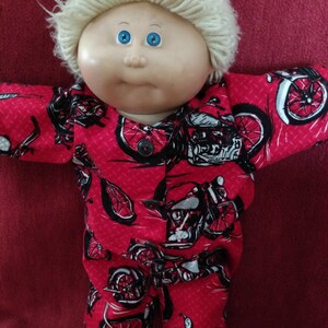 Könnte beinhalten: Eine Cabbage Patch Kid Puppe, die ein rot-schwarzes Schlafanzug-Outfit mit einem Motorrad-Druck trägt. Die Puppe hat blonde Haare und blaue Augen.