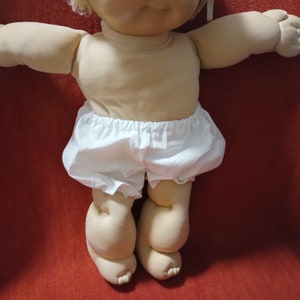 Puede incluir: Una muñeca Cabbage Patch Kid con cabello rubio y ojos azules que lleva un pañal blanco. La muñeca está sentada sobre una superficie roja.