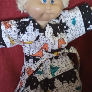 May include: A Cabbage Patch Kid doll wearing a black and white robe with a city skyline and dinosaur print. The doll has blonde hair and blue eyes.