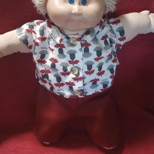 May include: A doll with light skin, blue eyes, and curly blonde yarn hair. The doll wears a white short-sleeved collared shirt featuring a repeating pattern of grey and red thistle illustrations with the word THISTLE, and solid dark red trousers.