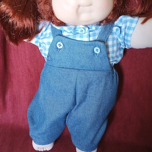May include: A Cabbage Patch Kid doll with red hair, wearing a blue denim overall with a white and blue gingham shirt.
