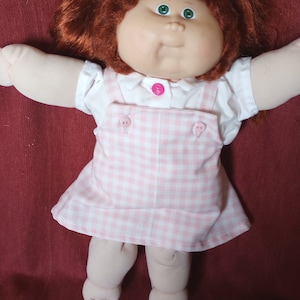 May include: A vintage doll with red hair styled in pigtails, green eyes, and a round face. The doll wears a white shirt and a pink and white checkered overall dress. The doll's arms and legs are extended, and it is set against a burgundy background.