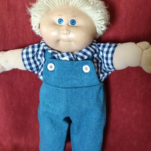 Puede incluir: Una muñeca Cabbage Patch Kid con cabello rubio, ojos azules y una camisa a cuadros azules y blancos. La muñeca lleva un mono azul y tiene una expresión dulce.