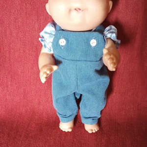 Peut inclure: Une poupée Cabbage Patch Kid portant une salopette en jean bleu et une chemise à carreaux blancs et bleus. La poupée a une crête brune et des yeux verts.