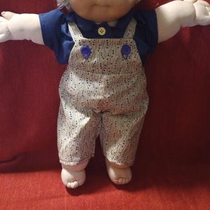 May include: A vintage doll with a soft, vinyl face, blue eyes, and a light blonde yarn hairstyle. The doll wears a navy blue shirt under patterned overalls with blue buttons. The doll's arms are outstretched, and it is posed against a red background.