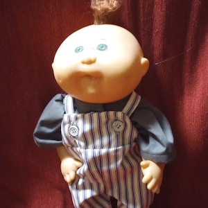 May include: A Cabbage Patch Kid doll with a red and white striped overall and a gray shirt. The doll has a bald head with a brown hairpiece and green eyes.