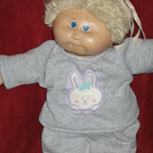 May include: A Cabbage Patch Kid doll wearing a gray and white pajama set with a bunny applique. The doll has blonde hair and blue eyes.