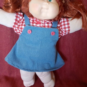 May include: A Cabbage Patch Kid doll with red hair, wearing a red and white gingham shirt and blue overalls. The doll has green eyes and a big smile.