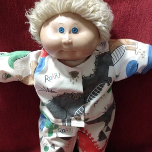 May include: A Cabbage Patch Kid doll wearing a white and blue dinosaur print pajama set with a white furry hat. The pajamas have the word "Roar!" printed on them.