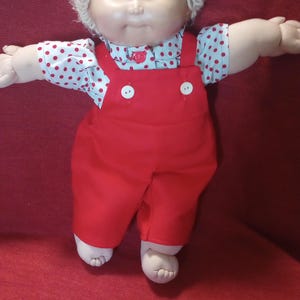May include: A doll with light skin, blue eyes, and short blonde yarn hair. The doll wears a white shirt with small red polka dots and bright red overalls featuring two white buttons on the front chest area. The doll has outstretched arms.