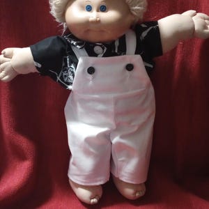 Puede incluir: Muñeca Cabbage Patch Kid vintage con una camisa negra con un diseño gráfico y un mono blanco. La muñeca tiene tez clara, ojos azules y un peinado rubio y esponjoso. El mono tiene dos botones negros.