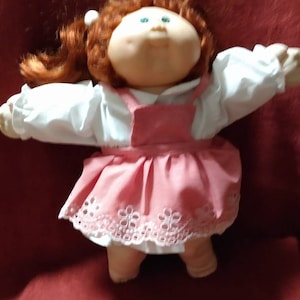 May include: A Cabbage Patch Kid doll with red hair, wearing a white blouse and a pink dress with white lace trim.