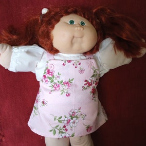 May include: A Cabbage Patch Kid doll with red hair wearing a pink floral dress with white buttons.