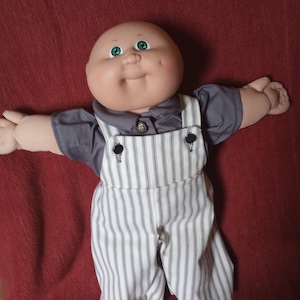 May include: A bald Cabbage Patch Kid doll wearing a grey shirt and white and black striped overalls. The doll has green eyes and rosy cheeks.