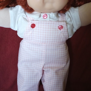 Op de afbeelding: Een Cabbage Patch Kid pop met rood haar, die een wit shirt en een roze-witte gingham-overall draagt.