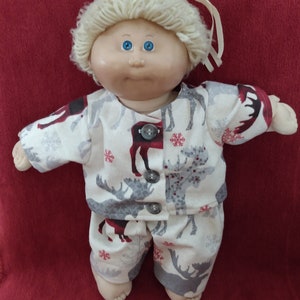 May include: A Cabbage Patch Kid doll wearing a white and gray flannel pajama set with a red and gray moose and snowflake print. The doll has blonde hair and blue eyes.