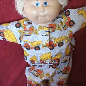 May include: A Cabbage Patch Kid doll with blonde hair wearing a grey and white pyjama onesie with a yellow and orange construction vehicle print.