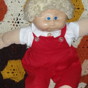 May include: A Cabbage Patch Kid doll with blonde hair wearing a white shirt and red dungarees. The doll is lying on a crocheted blanket with a variety of colours.