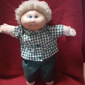 May include: A doll with a round face, bright blue eyes, and short, curly blonde yarn hair. The doll wears a green and white gingham button-down shirt with a collar and solid dark green pants. The doll has outstretched arms and a soft body.