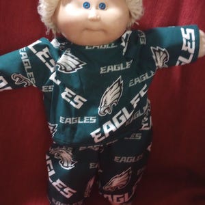 May include: A doll with a round face, blue eyes, and short blonde yarn hair. The doll wears a dark green long-sleeved shirt and matching pants featuring a repeating white pattern of the Philadelphia Eagles logo and the text EAGLES.