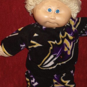 May include: A Cabbage Patch Kid doll wearing a black, purple, and yellow fleece pajama set with a graphic print of a stylized animal.
