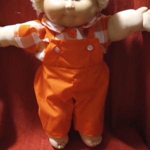 Pode incluir: Uma boneca vintage com um rosto redondo, olhos azuis e um penteado loiro fofo. A boneca usa um macacão laranja com botões brancos e uma camisa xadrez. Os braços da boneca estão estendidos.