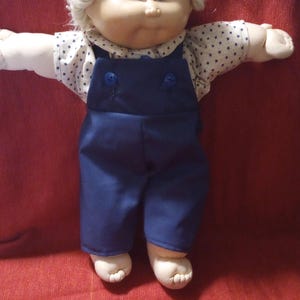 May include: A vintage doll with a soft, round face, blue eyes, and a fluffy, light-coloured hairstyle. The doll is wearing a white shirt with a blue star pattern and blue dungarees. The doll's arms are outstretched.