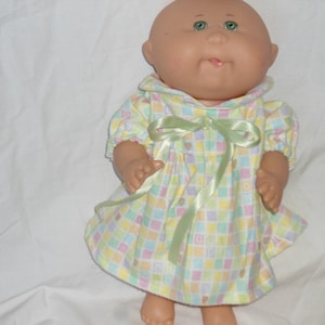 May include: A Cabbage Patch Kid doll wearing a light green, yellow, pink, and blue checkered dress with a light green satin ribbon tied at the waist.