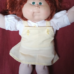 May include: A Cabbage Patch Kid doll with red hair, green eyes, and a white shirt with yellow overalls. The doll is lying on a red surface.