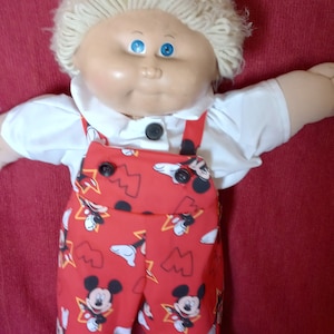 May include: A Cabbage Patch Kid doll wearing a white shirt and red overalls with a Mickey Mouse print. The doll has blonde hair and blue eyes.