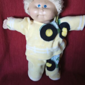 May include: A Cabbage Patch Kid doll wearing a yellow fleece pajama set with a tractor print. The doll has blonde hair and blue eyes.